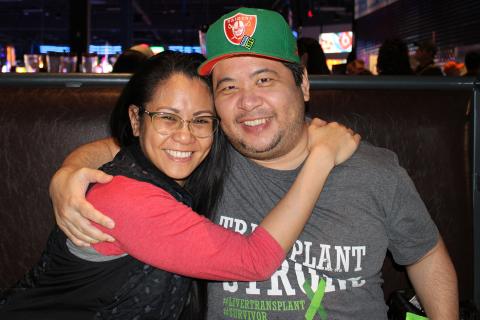 Angelo and a woman sit in a booth together, arms around each other, smiling at the camera.